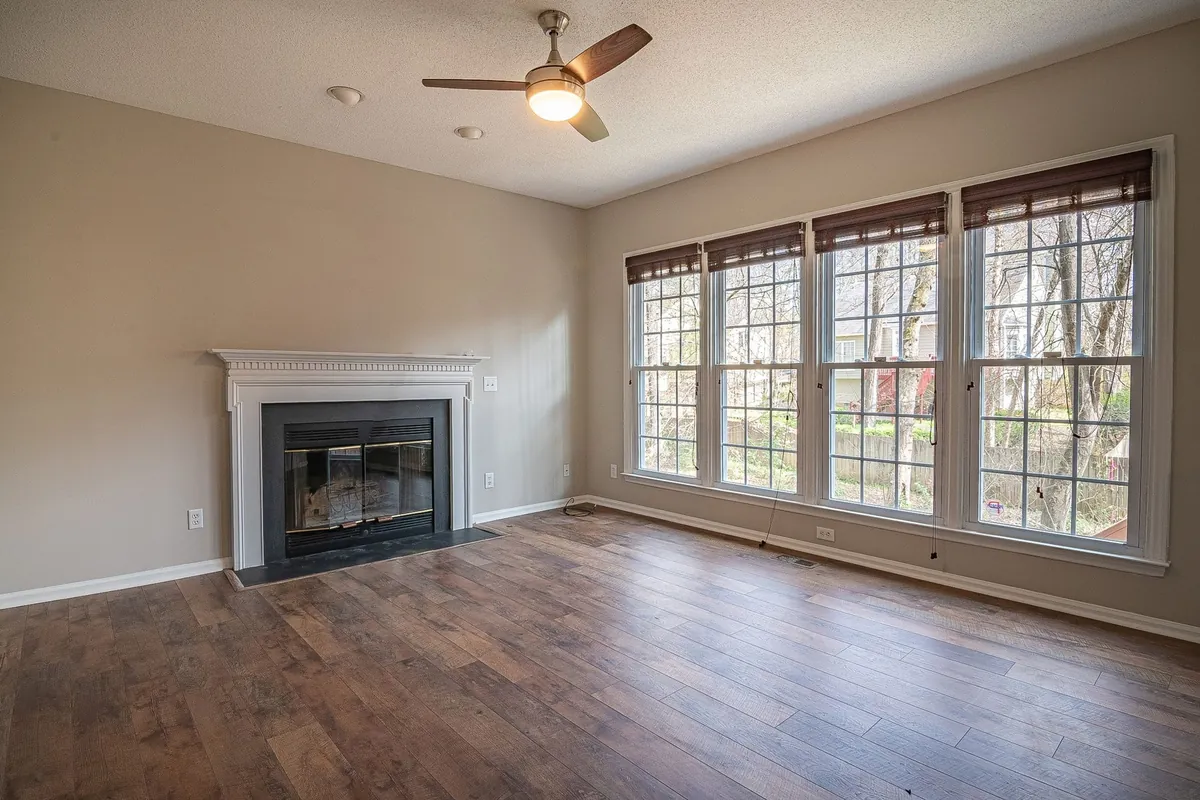 An empty living room with a white fireplace mantel, a wall of four tall windows with wood blinds, a ceiling fan and a dark wood floor